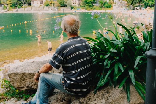 Senior man enjoying leisure time by a serene lake surrounded by nature and people swimming.