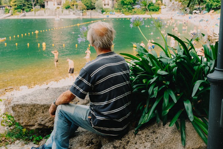 Elderly Man In Striped Shirt Sitting On Rock Watching People On The Lake