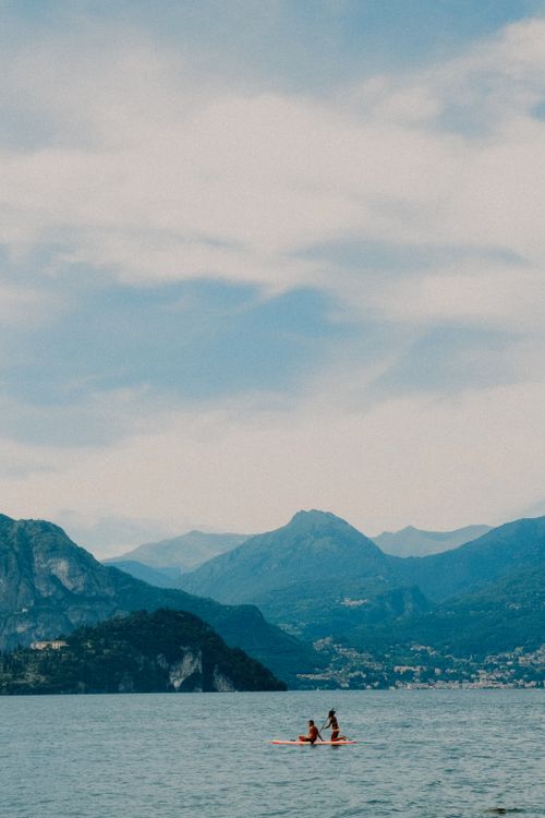 Free Tranquil scene of a lake with people paddleboarding and mountain views under a cloudy sky. Stock Photo