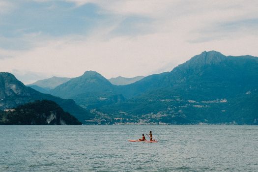 Two people kayaking on a tranquil lake surrounded by scenic mountains under a cloudy sky.