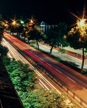 High angle shot of a highway at night with dynamic light trails amidst lush trees and glowing streetlights.