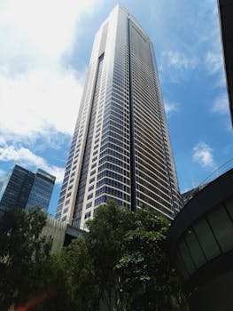 Tall skyscraper with modern architecture reaching into a clear blue sky, surrounded by urban greenery.