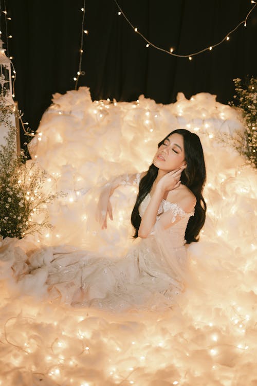 Woman in White Dress Sitting Beside Fairy Lights