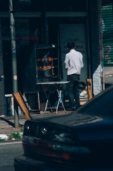 Man roasting meat at a street vendor near a parked black car in an urban setting.