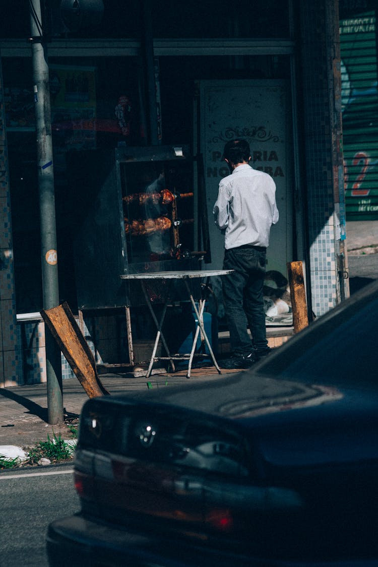 Man Roasting Chicken On A Street And Black Car Parked In Foreground