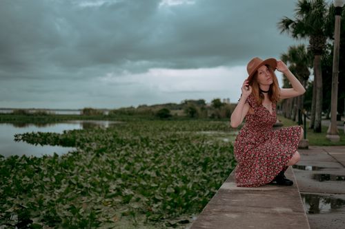 Free A woman in a floral dress and hat poses by a lush lakeside on an overcast day. Stock Photo