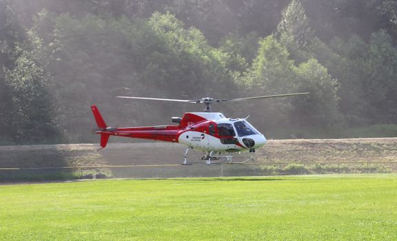 Aerial view of a red and white helicopter flying above a green field against a forest backdrop.
