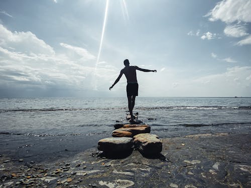 Free A Silhouette of a Man Balancing on Stones at a Shore Stock Photo