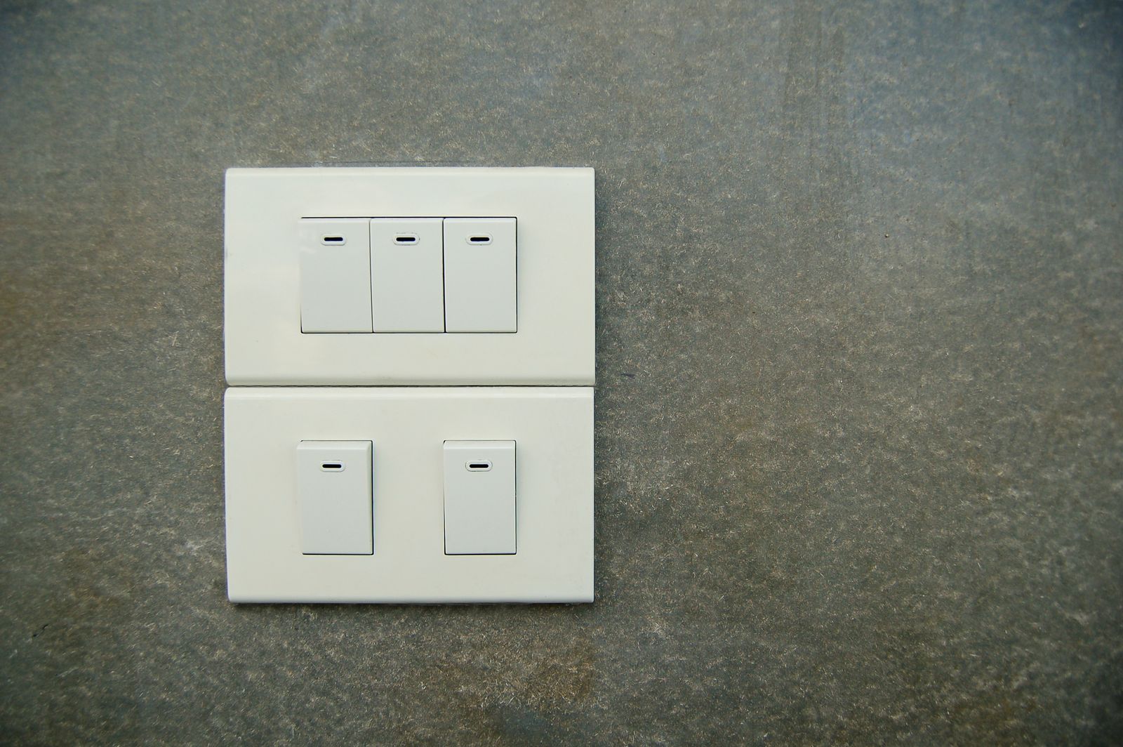 Two white rocker light switch plates mounted on a mottled grey stone wall, five switches in total, all in the off position
