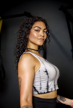 Portrait of a confident woman with curly hair, tattoos, and nose piercings in a gym setting.