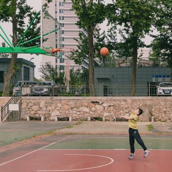 Young boy shooting a basketball on an outdoor court with urban buildings in the background.