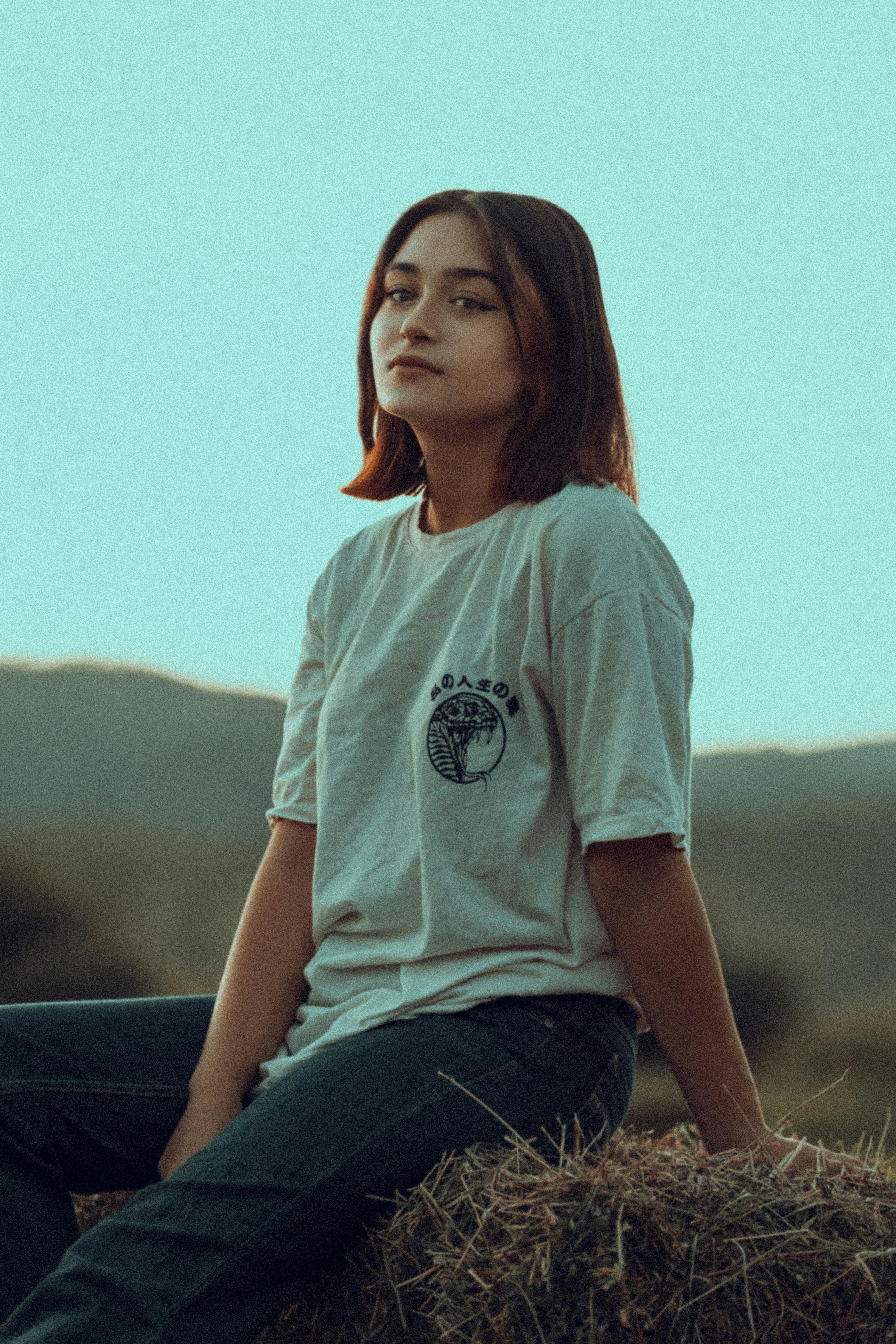 A young woman seated on hay bales outdoors wearing a casual t-shirt, exuding a relaxed vibe.