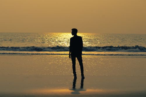 Free Silhouetted figure stands on a beach reflecting golden sunset hues with the ocean in the background. Stock Photo
