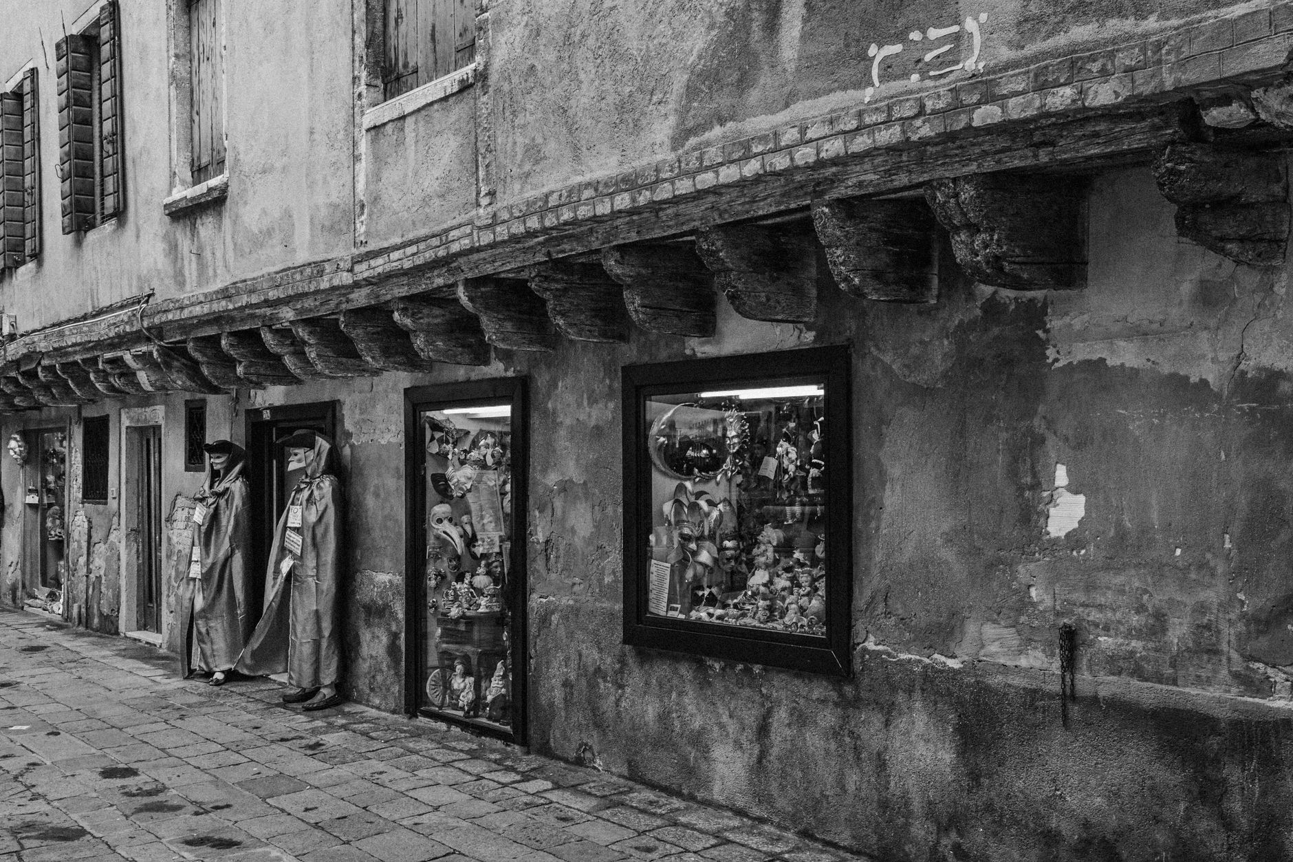Black and white photo of a Venetian street featuring carnival masks in shop and two mannequins.