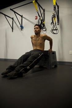 A shirtless man works out indoors, performing triceps dips in a gym setting.
