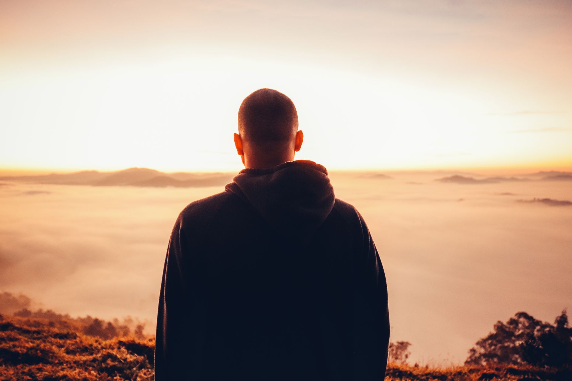 A solitary man in a hoodie stands gazing at a serene sunrise above a sea of clouds.
