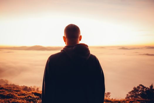 A solitary man in a hoodie stands gazing at a serene sunrise above a sea of clouds.