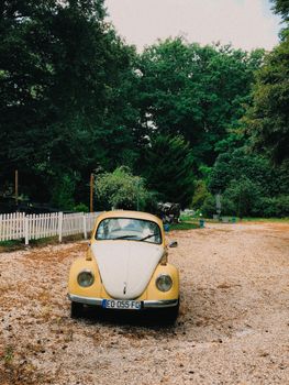 Classic yellow Volkswagen Beetle parked on a scenic dirt road with trees and a picket fence.