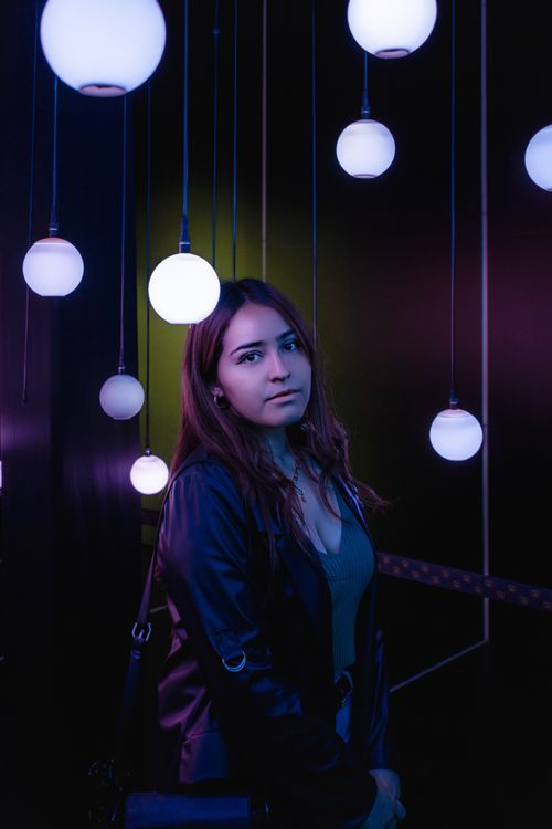 Free A woman in a jacket stands under glowing pendant lamps, creating a moody ambiance. Stock Photo