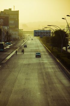 Early morning urban street scene with a few people crossing and cars during sunrise.