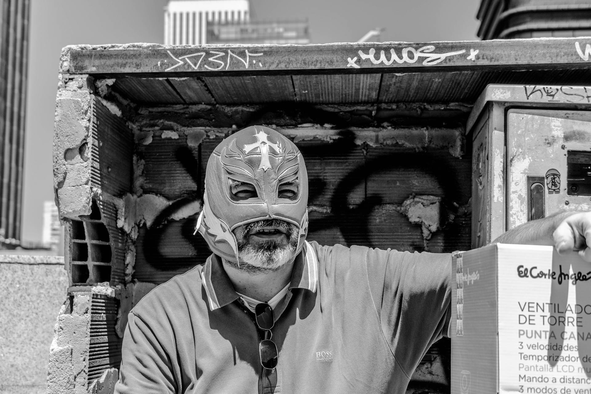 A man wearing a luchador mask poses with a box in a gritty urban environment.