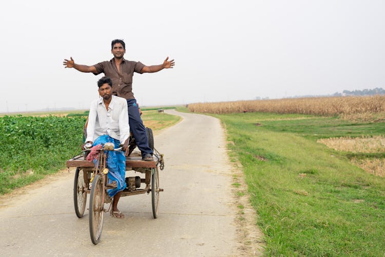 Men Riding A Bike Carriage On Road Between Green Grass