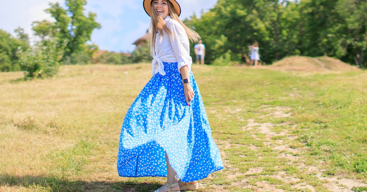 Woman with hat and blue skirt smiling in a sunny park setting. Summer vibes.
