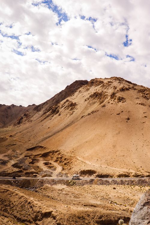 Free A car drives along a mountain road under a cloudy sky in a remote countryside landscape. Stock Photo