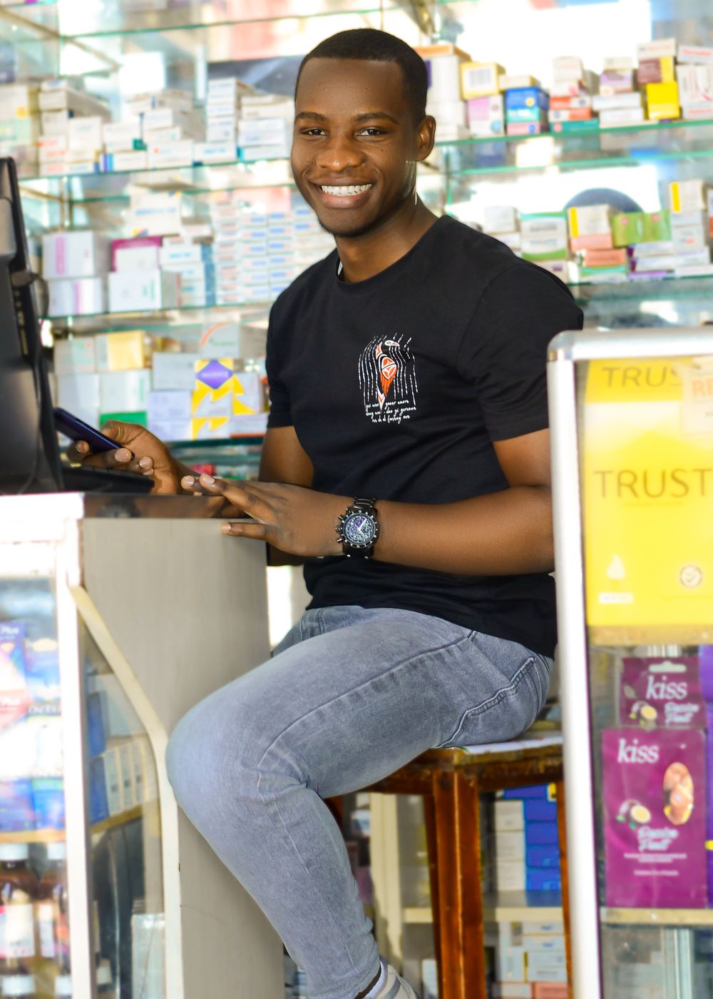 Smiling attendant behind the counter of a neighbourhood pharmacy, shelves of medicine behind him