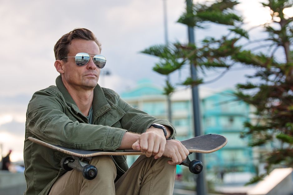 Casual man with sunglasses sitting at skate park, reflecting and relaxing with skateboard.