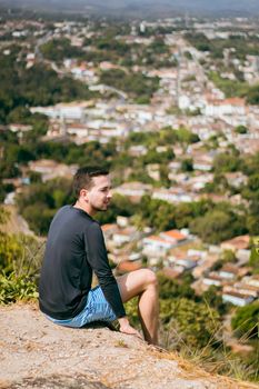 Man in striped shorts sitting on mountain overlooking Brazilian cityscape.