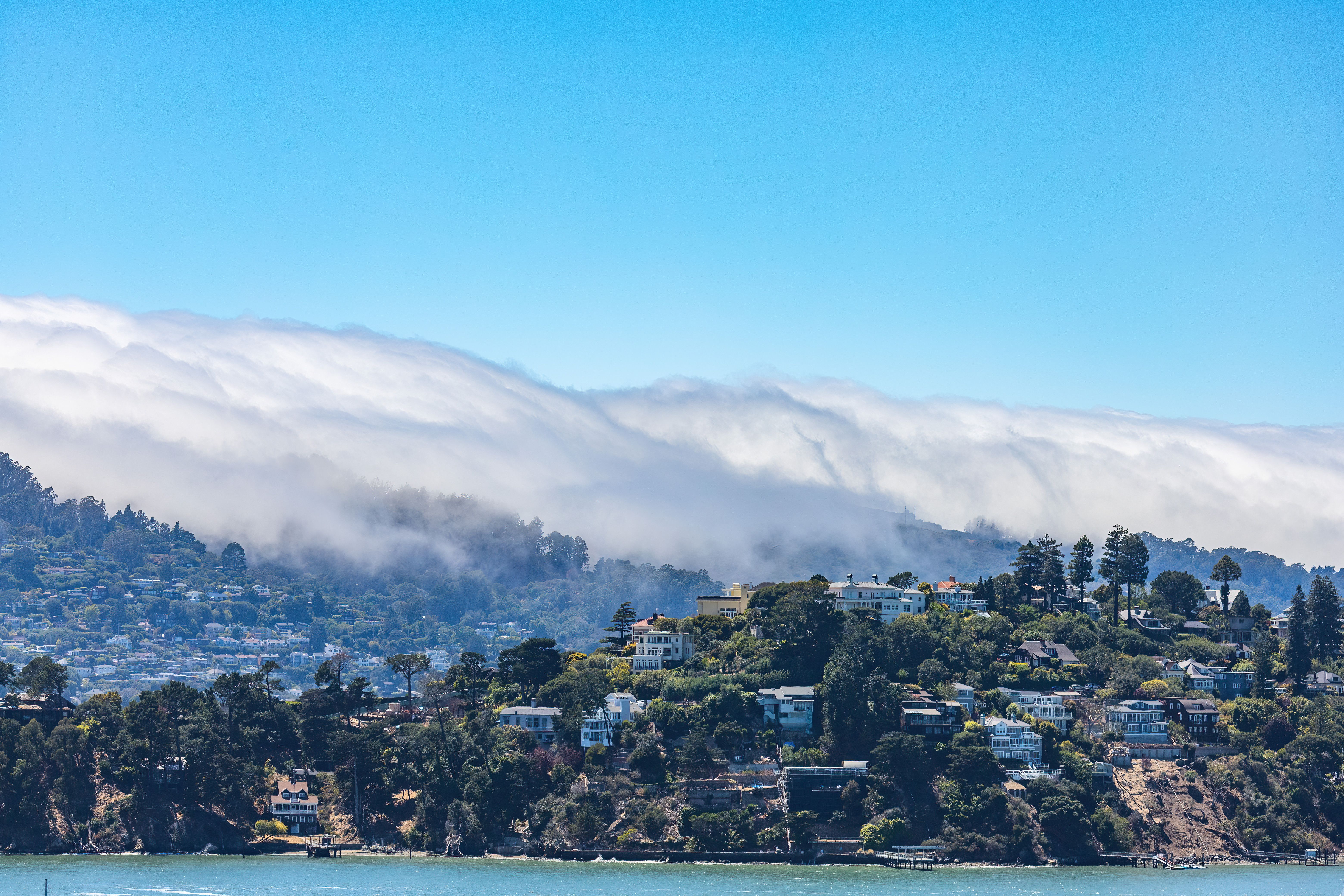 A scenic view of fog rolling over hills with houses by the waterfront, under a clear sky.