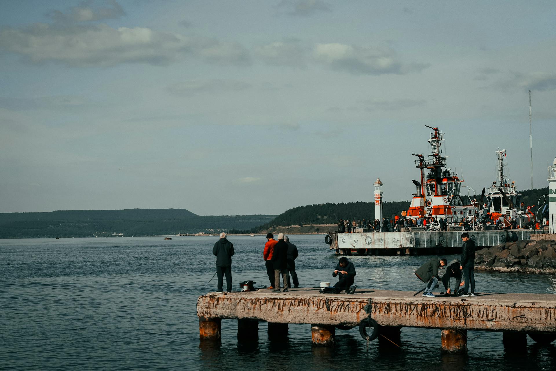 A scenic view of a harbor with people fishing on a pier and boats in the background.