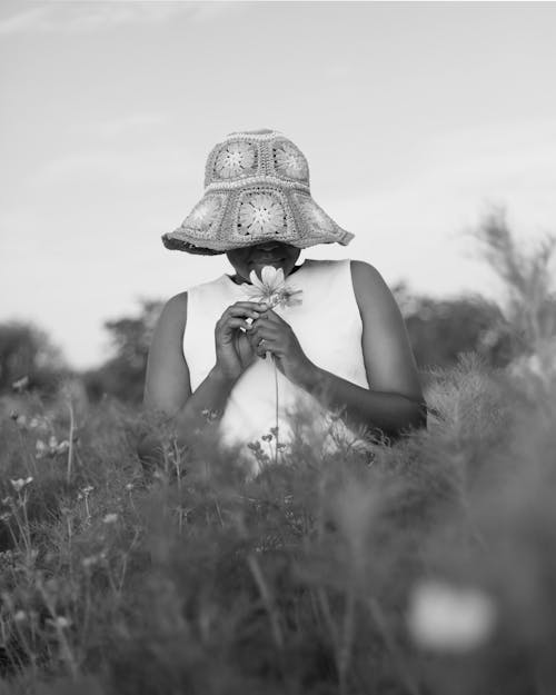 Grayscale Photo of Woman Face Covered with Knitted Hat Standing on a Field Holding Flower