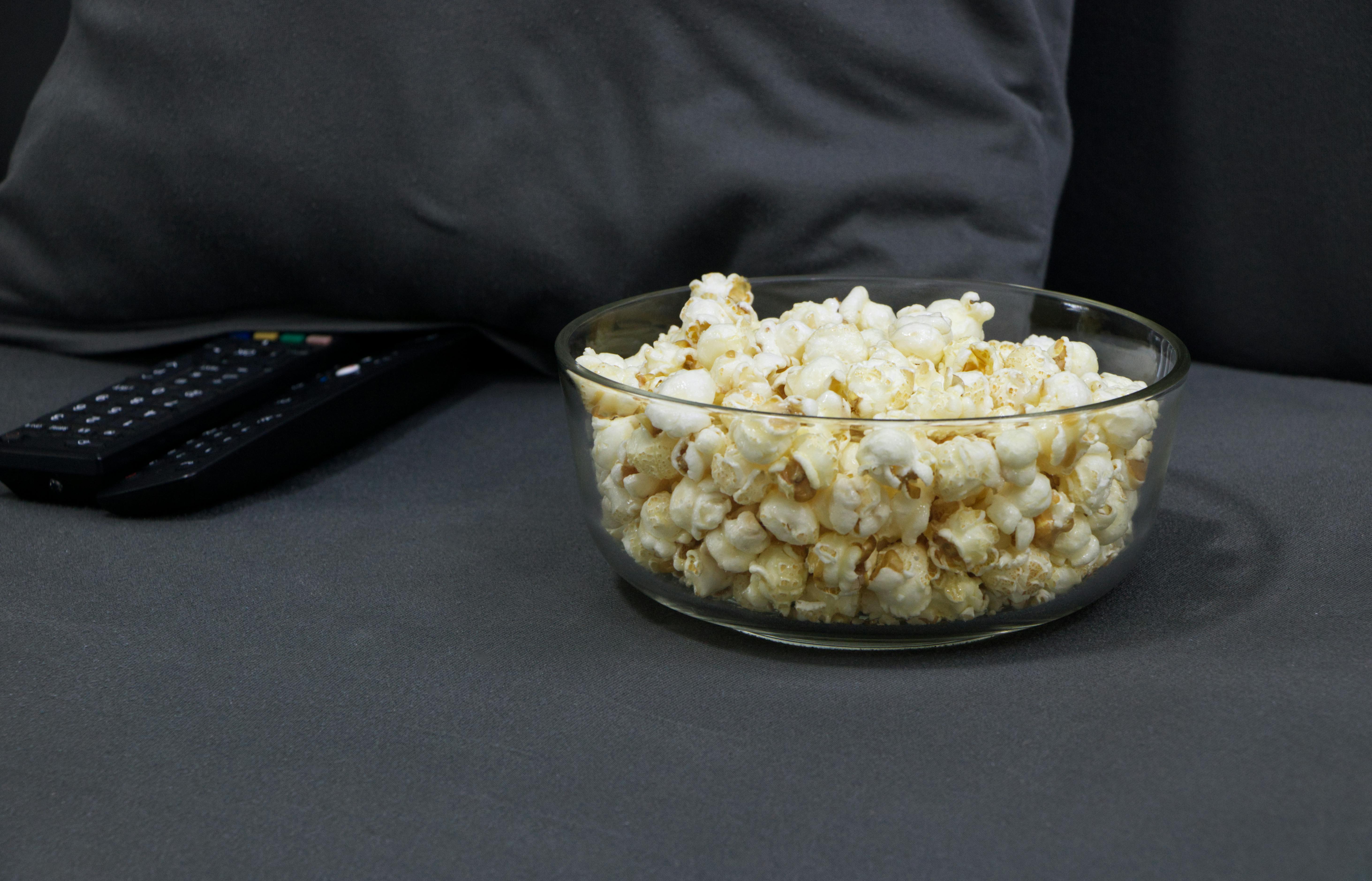 Glass bowl filled with popcorn next to TV remotes on a gray sofa. Perfect for cozy movie nights.