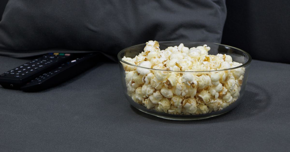 Glass bowl filled with popcorn next to TV remotes on a gray sofa. Perfect for cozy movie nights.
