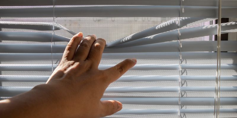 Person opening white window blinds to let sunlight in