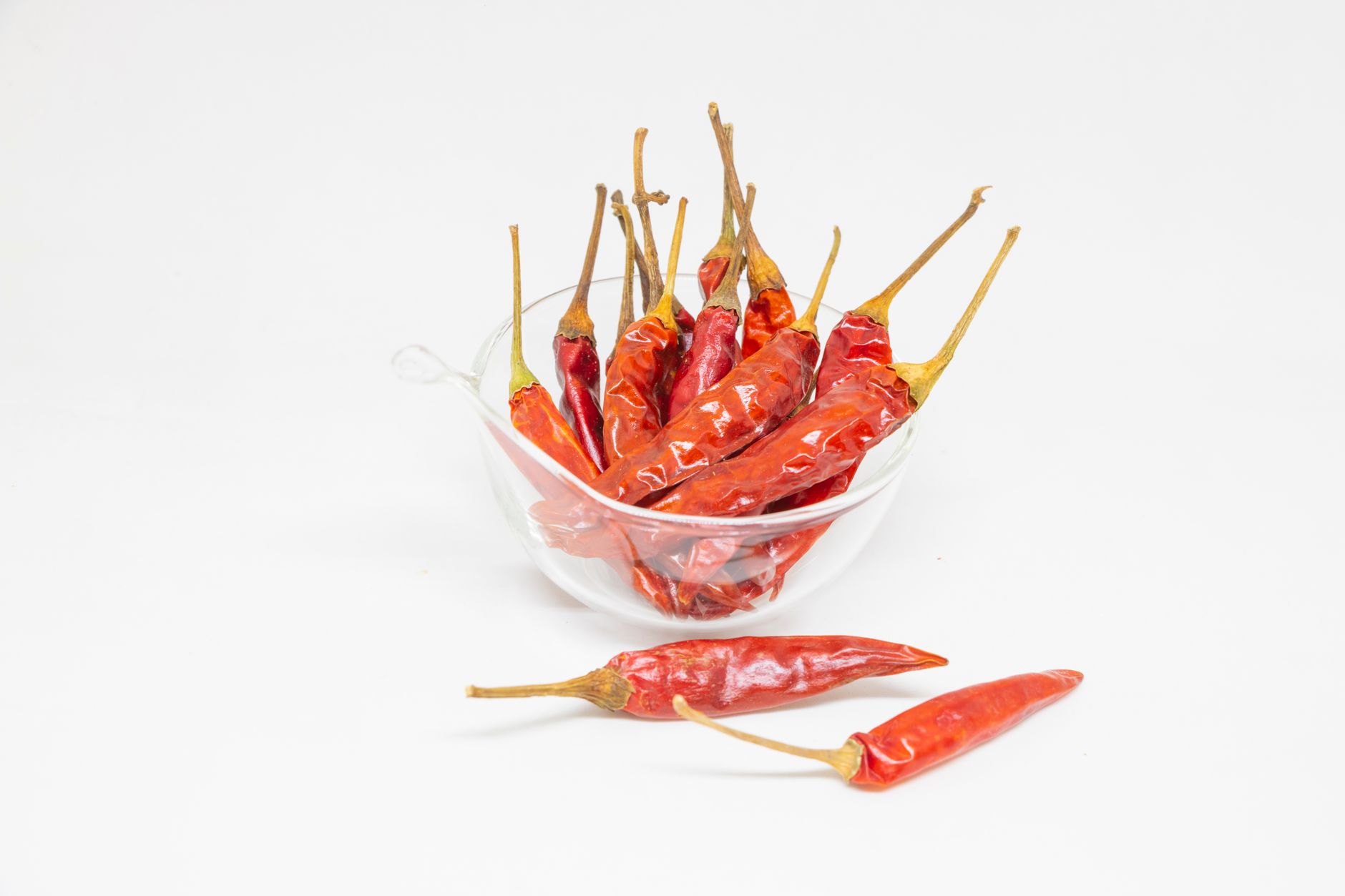 A close-up of dried red chillies in a glass bowl on a white background, showcasing vibrant spices.