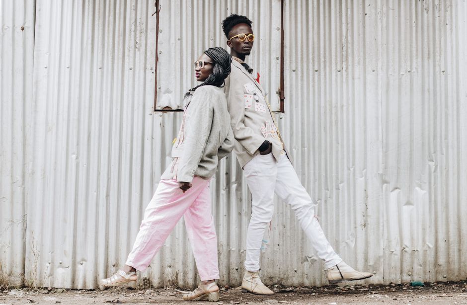 Two African models in stylish, vintage fashion leaning against a corrugated wall outdoors.