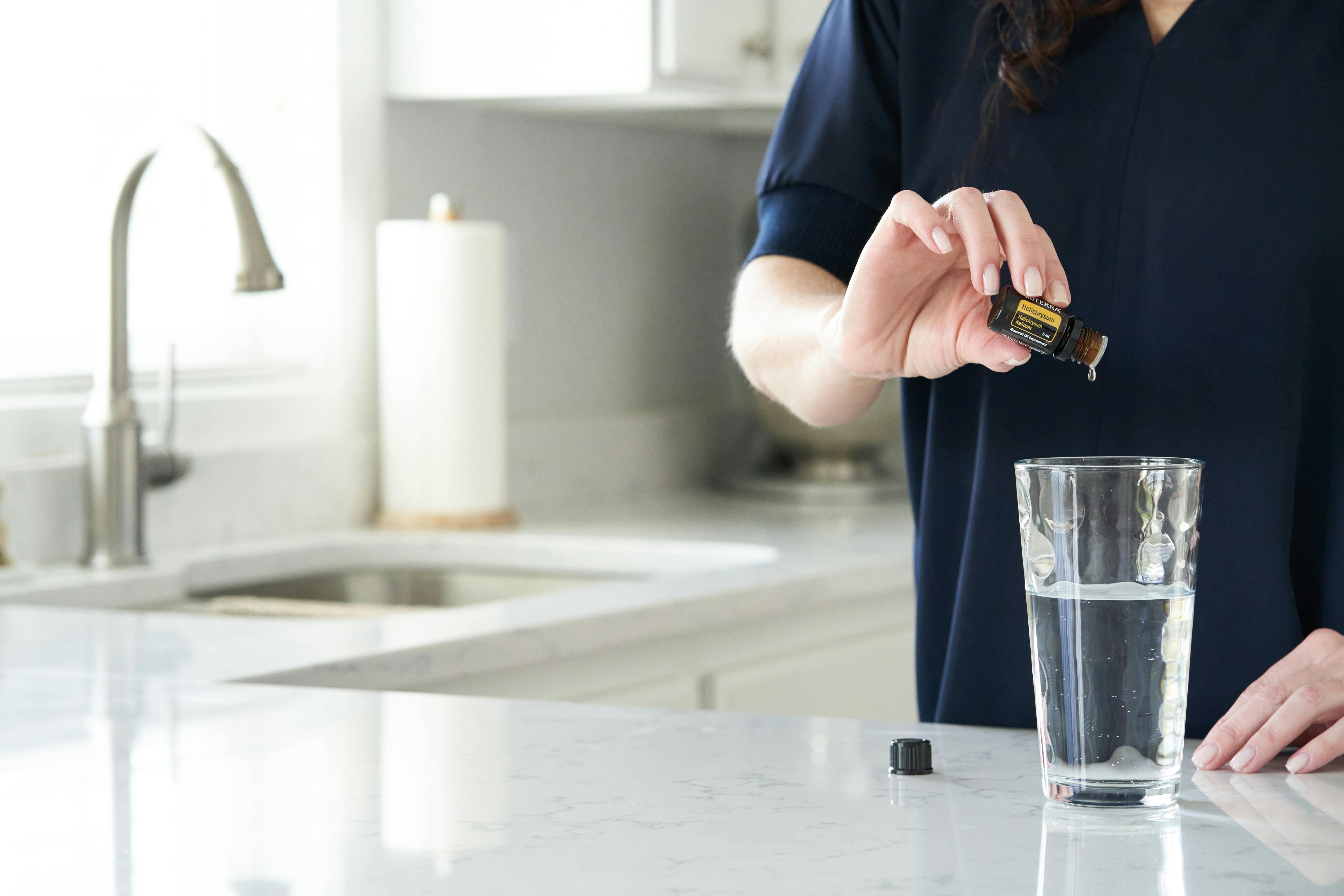 Person pouring essential oil into a glass of water on a kitchen countertop.