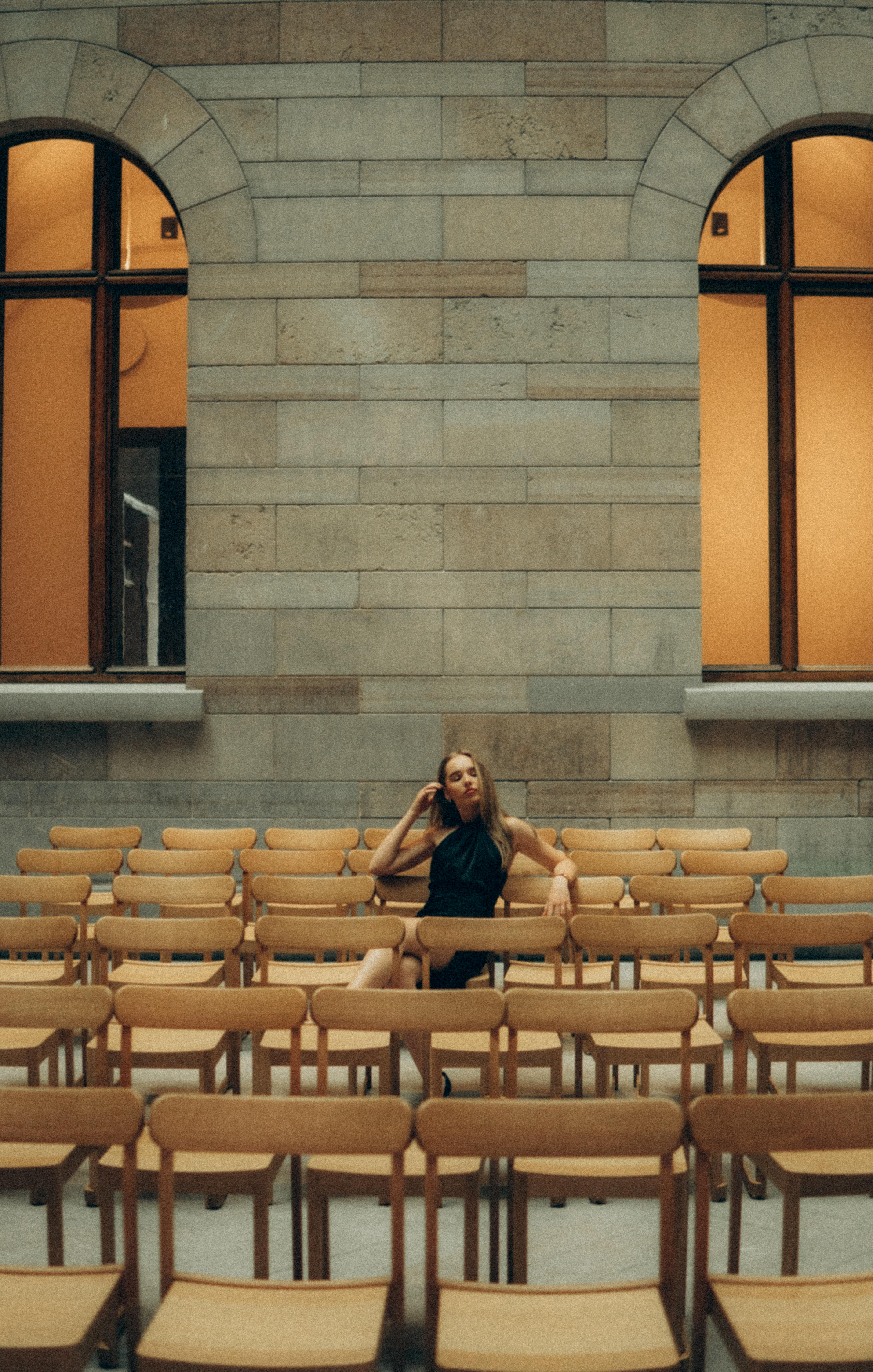 A stylish woman sitting alone among neat rows of empty wooden chairs indoors.