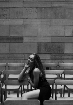 A woman in a black dress poses thoughtfully sitting among rows of chairs in a serene monochrome setting.