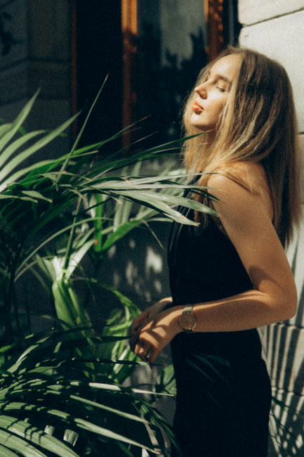 A woman in a black dress enjoying sunlight amidst vibrant green plants, creates a serene and stylish scene.