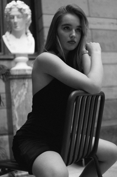 Stylish woman in a sleeveless dress poses seated indoors in grayscale setting.