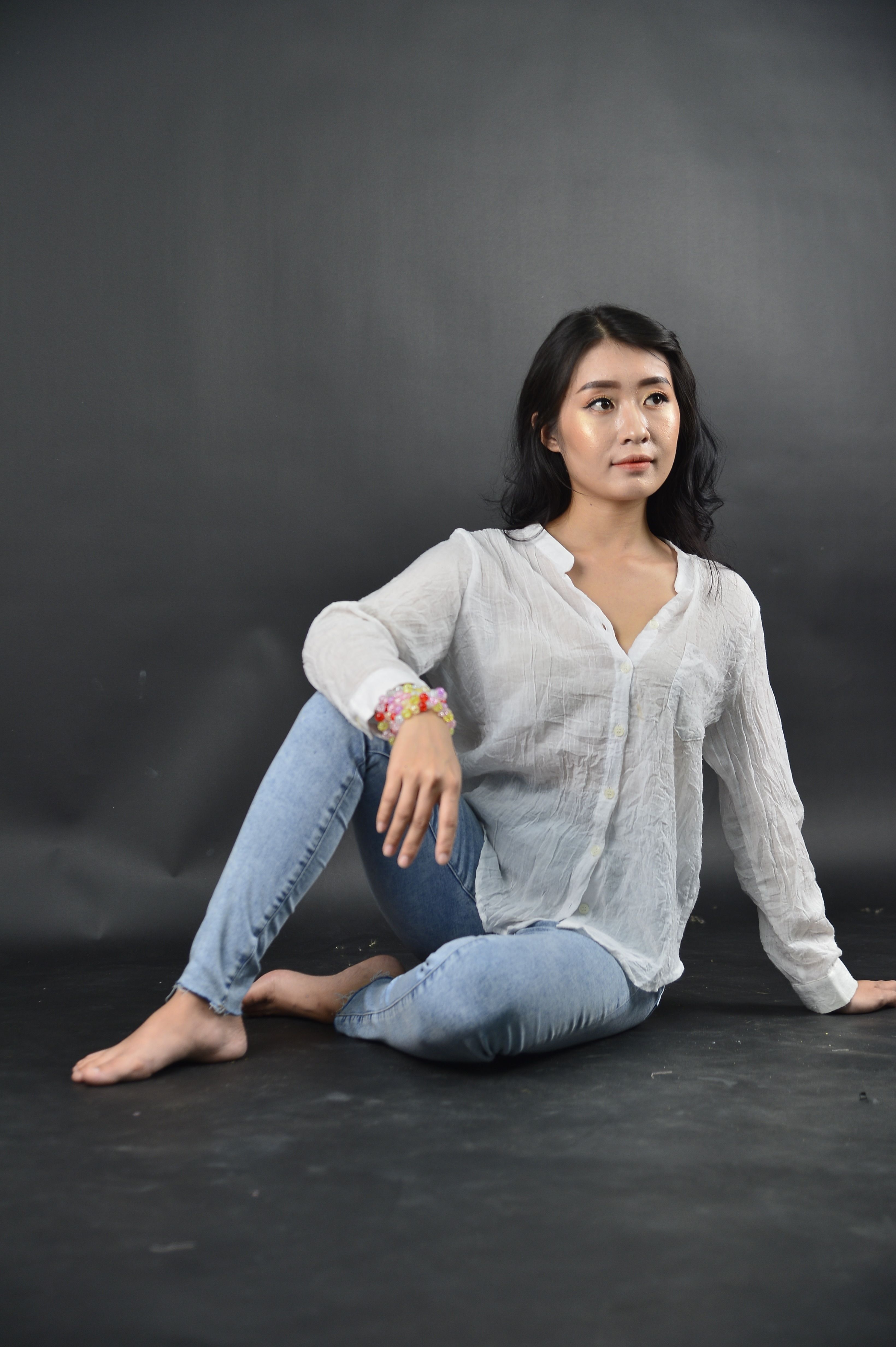 Studio portrait of an Asian woman sitting, wearing denim pants and white long sleeves.