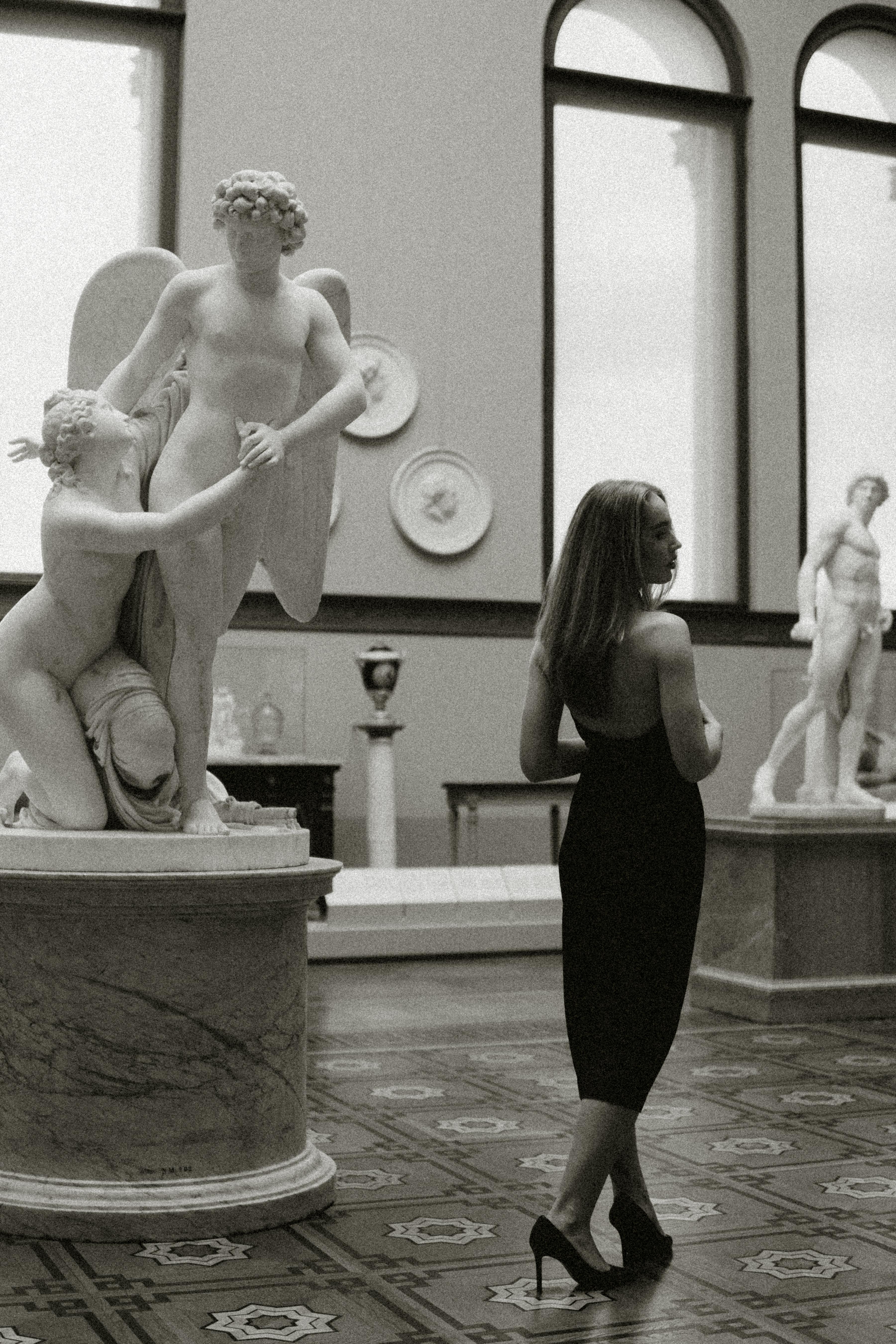 Free Monochrome image of an elegant woman standing among classical sculptures in a museum. Stock Photo