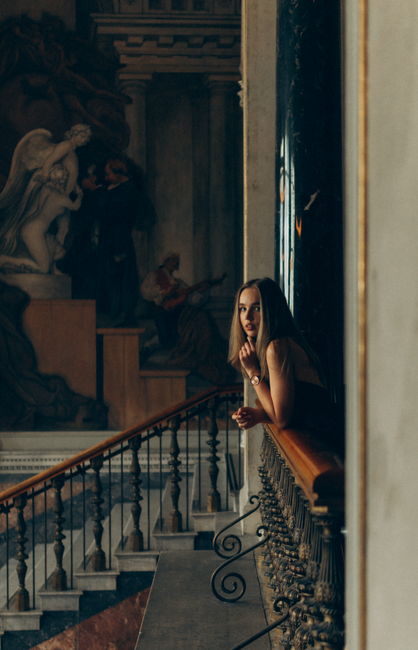A woman leans on a railing in a museum, gazing contemplatively at art pieces with a serene demeanor.