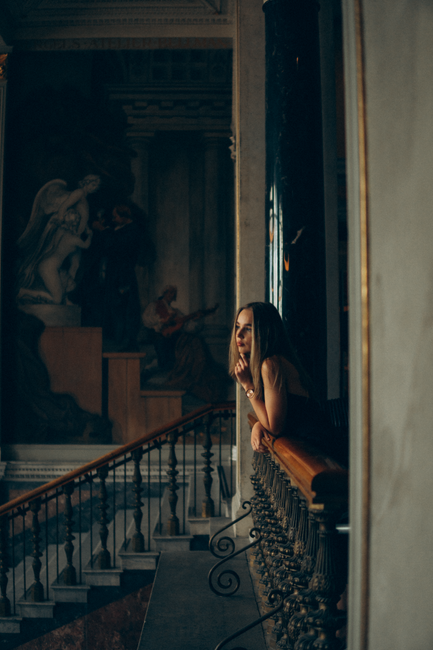 A woman leans on a railing, pondering in an elegant, dimly lit museum.