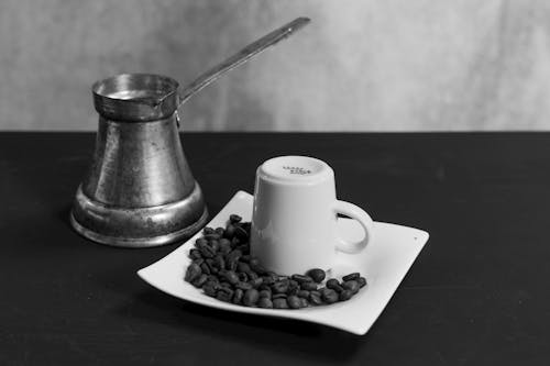 A Grayscale of a Cezve beside a Cup and a Saucer with Coffee Beans