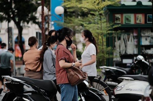 A Woman Wearing Black Face Mask Standing Near People Sitting on Motorbike